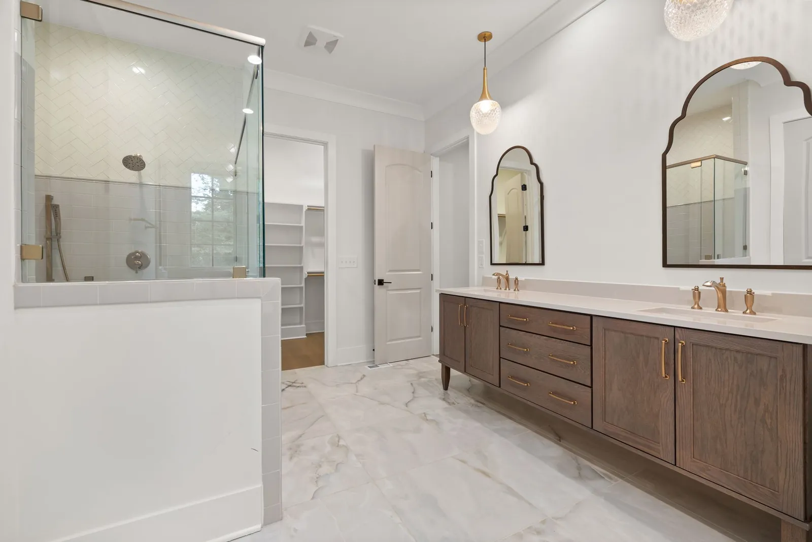 A bright bathroom with a wood vanity, arched mirrors and a glass shower